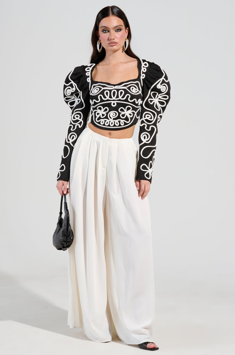A person stands against a plain background wearing the Blurred Lines Long Sleeve Blouse with puffed sleeves and high-waisted, wide-leg white trousers. They hold a small black handbag and wear black sandals.