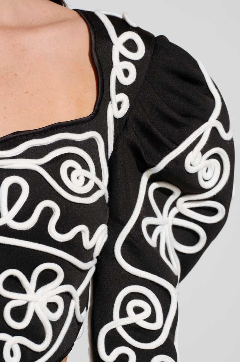 The BLURRED LINES LONG SLEEVE BLOUSE features a detailed close-up of its black cropped design, adorned with intricate white embroidered swirls and floral motifs. The textured fabric is accentuated by puffed sleeves, adding an elegant touch to the overall blouse design.