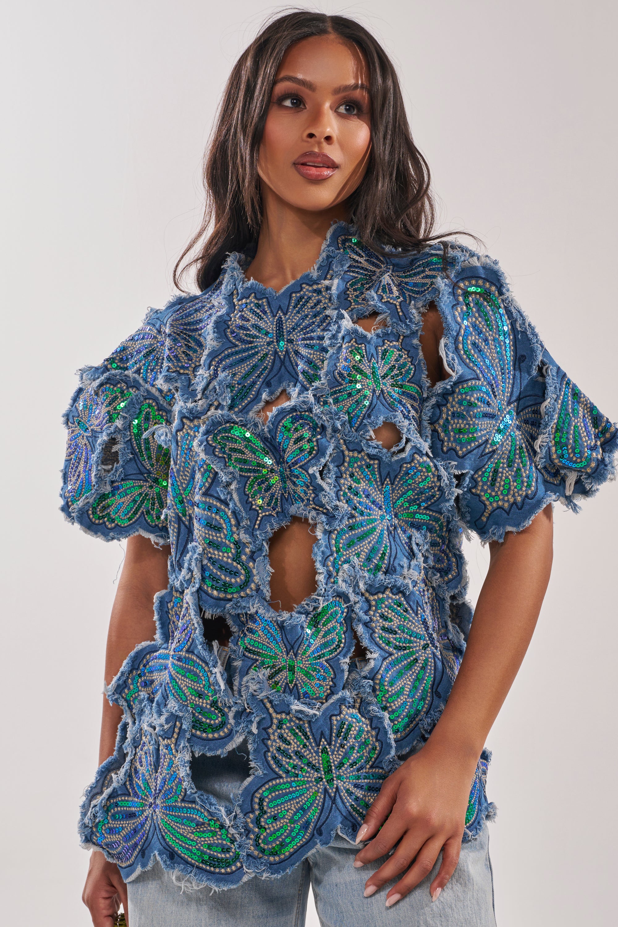A woman wears the SPREAD YOUR WINGS DENIM TOP—featuring large, intricate butterfly designs adorned with green and blue beading, cut-out sections, and frayed edges. Her confident look makes this a bold festival statement piece. 0