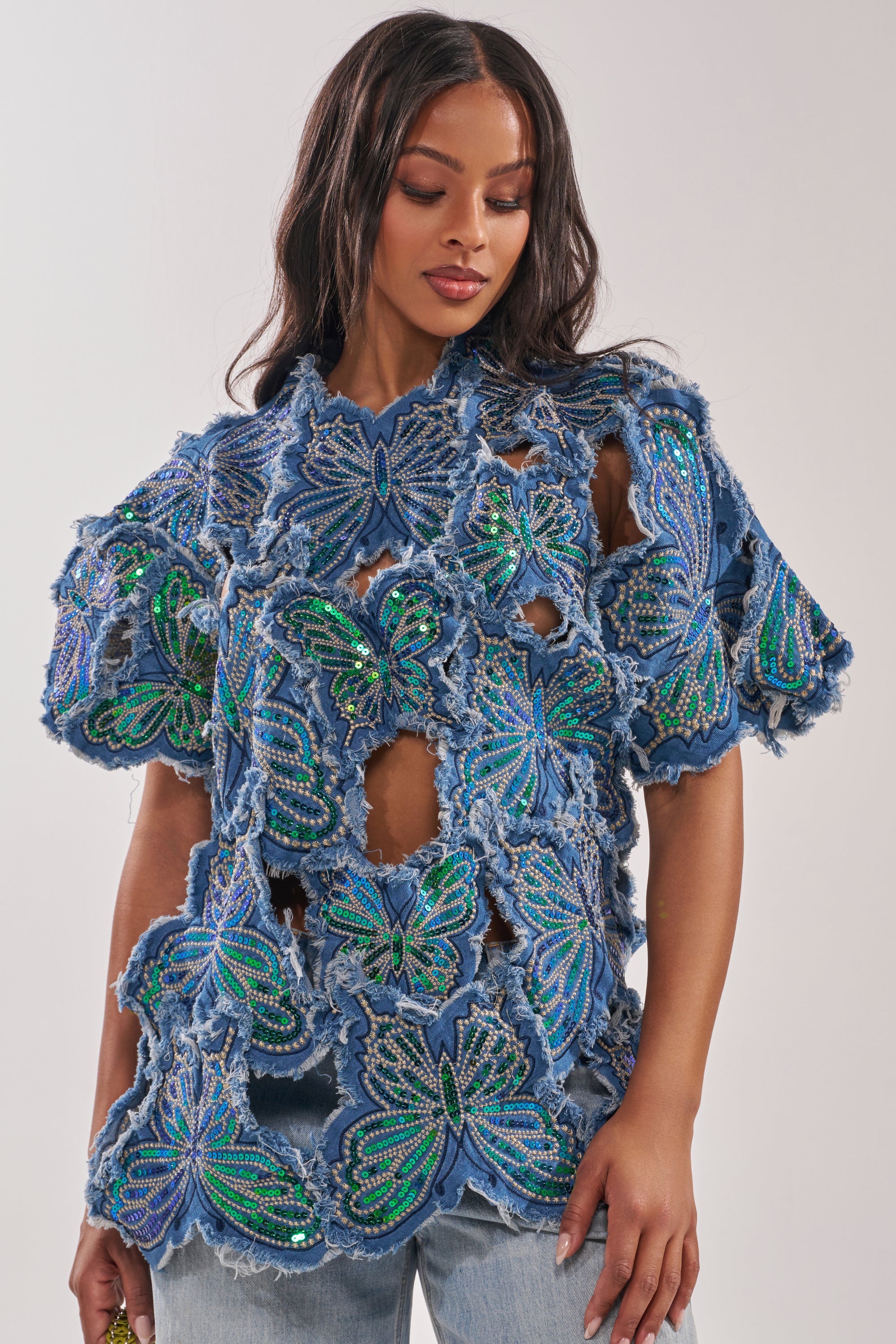 Wearing the SPREAD YOUR WINGS DENIM TOP with sequined butterfly accents and distressed edges over light blue jeans, a woman with wavy dark hair looks down, radiating effortless festival style. 3