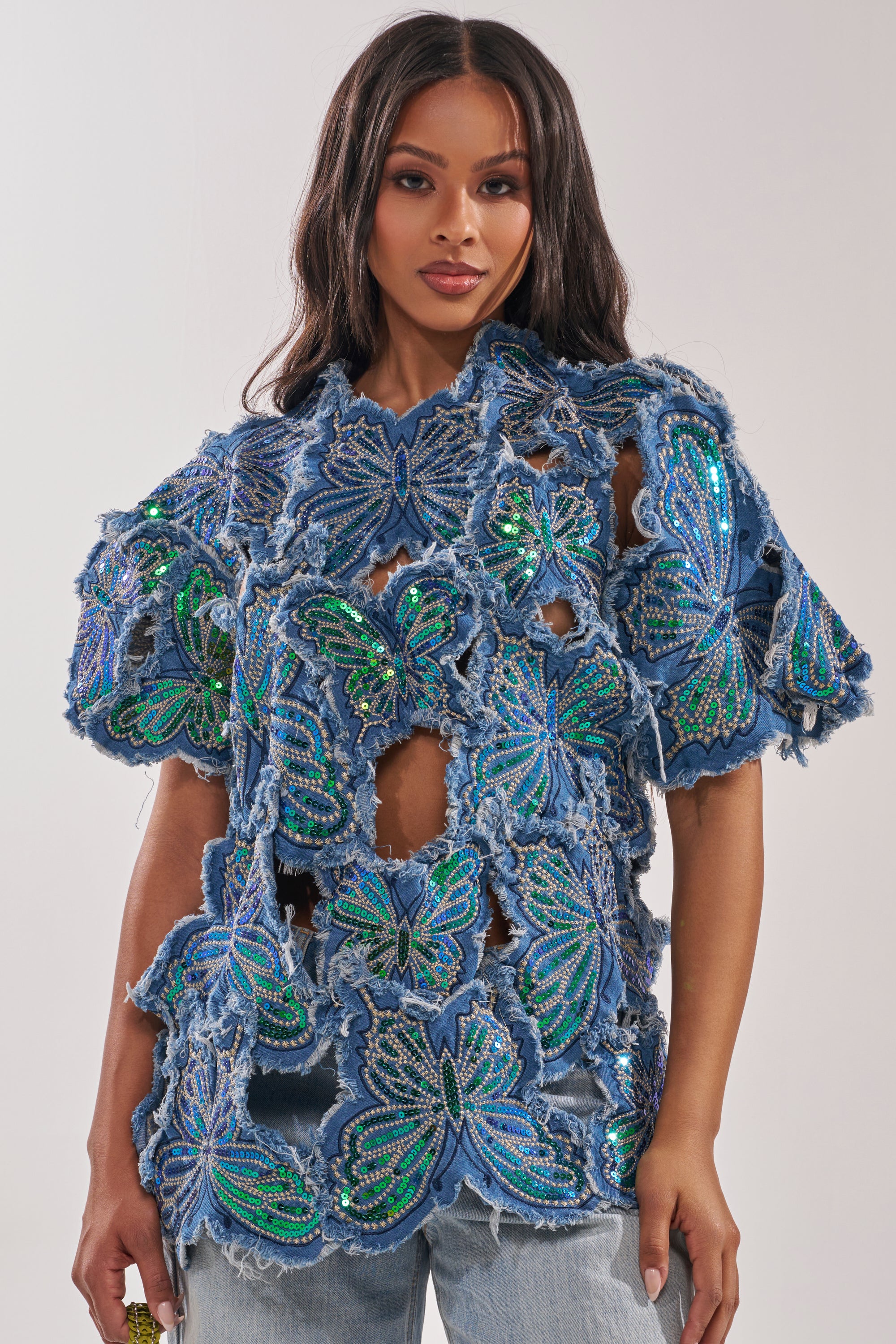 A woman models the SPREAD YOUR WINGS DENIM TOP, featuring large green and blue sequin butterflies and frayed edges, paired with light blue jeans—a festival-ready standout—posed against a plain background. 1