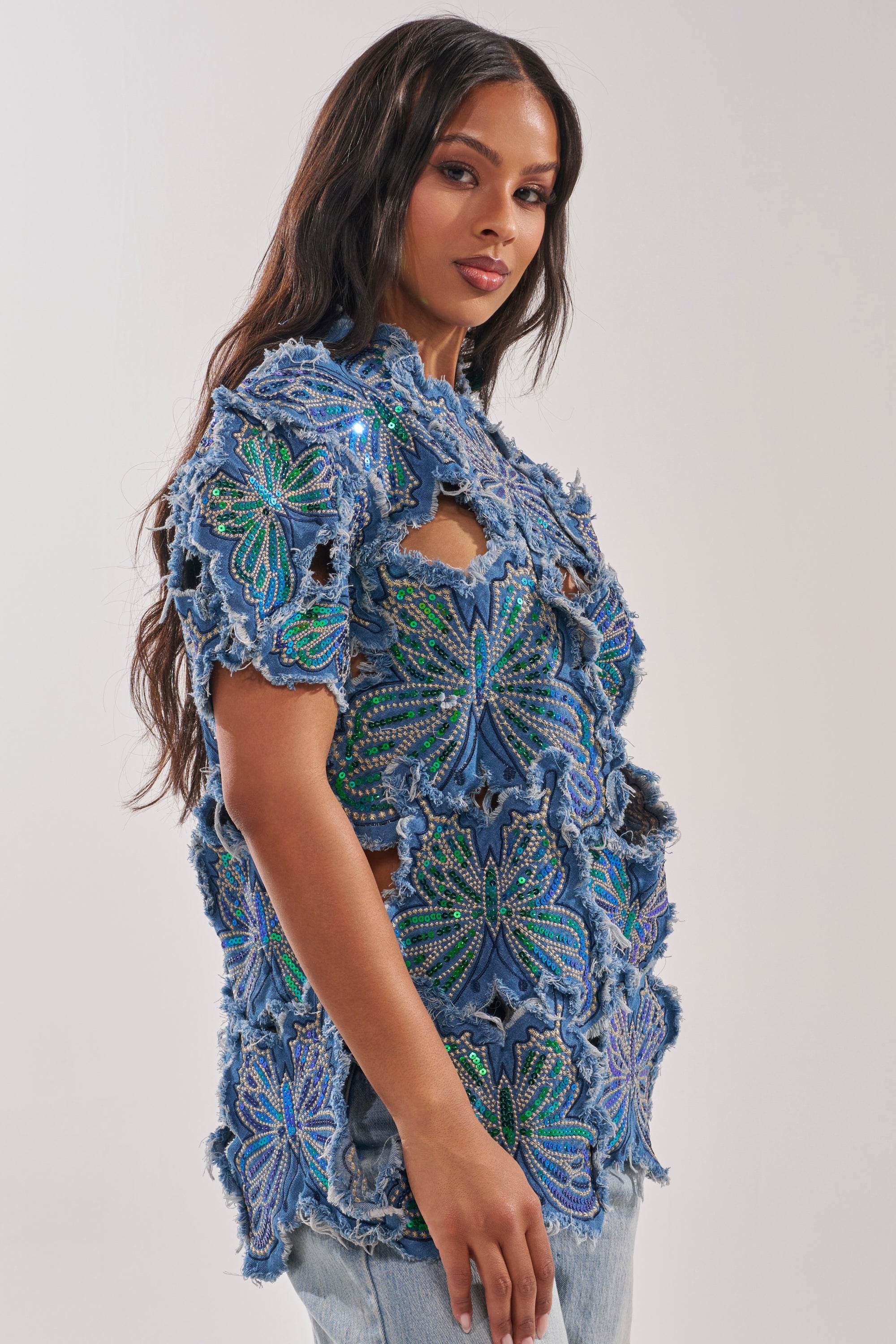 A woman with long dark hair models the SPREAD YOUR WINGS DENIM TOP, a blue rhinestone butterfly style with large beaded and embroidered floral patterns and cut-out details, paired with light denim jeans against a plain background. 2