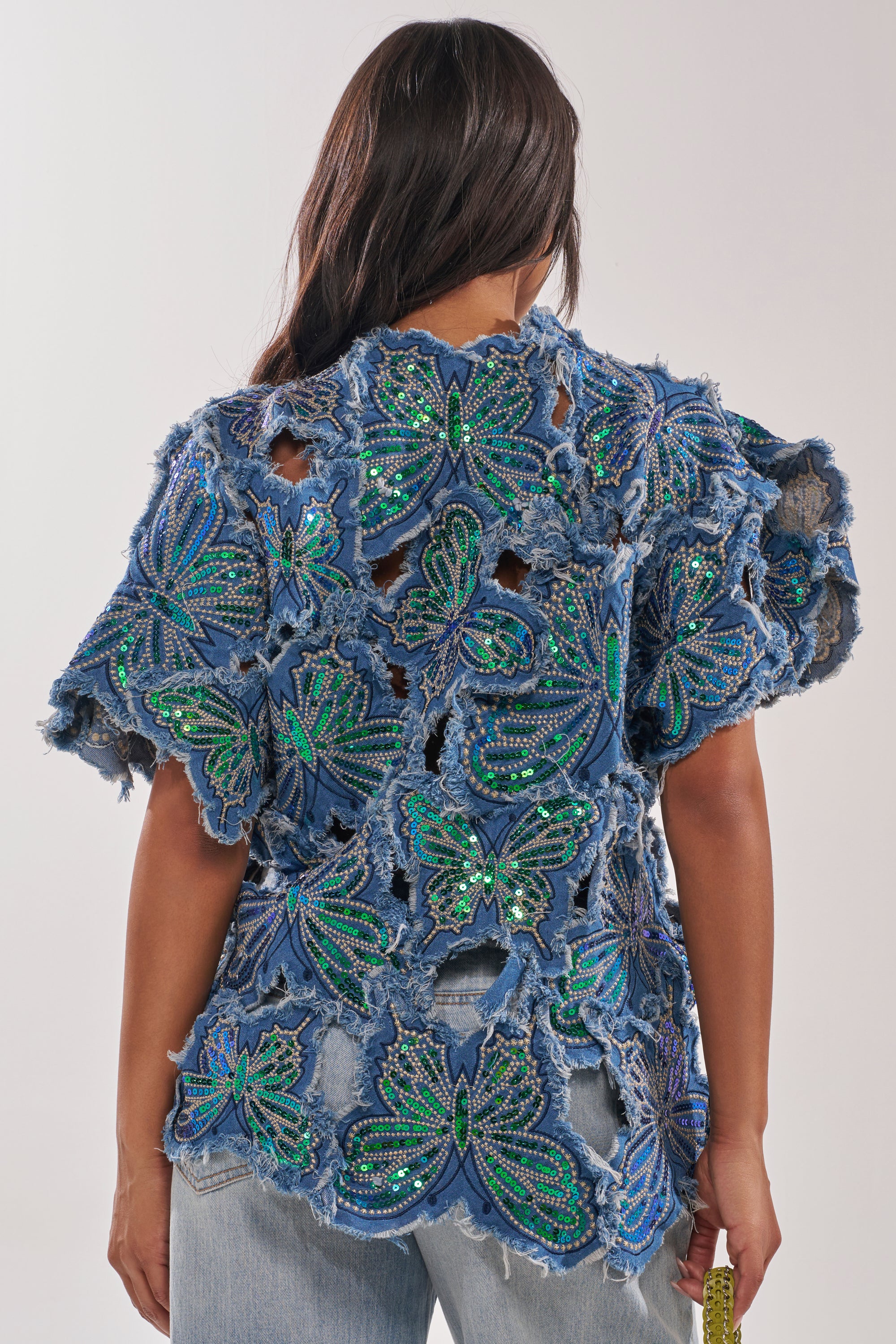 A person with long dark hair wears the SPREAD YOUR WINGS DENIM TOP, a blue rhinestone butterfly top with green and blue butterfly cutouts and distressed edges, styled from the back with light-wash jeans. 4