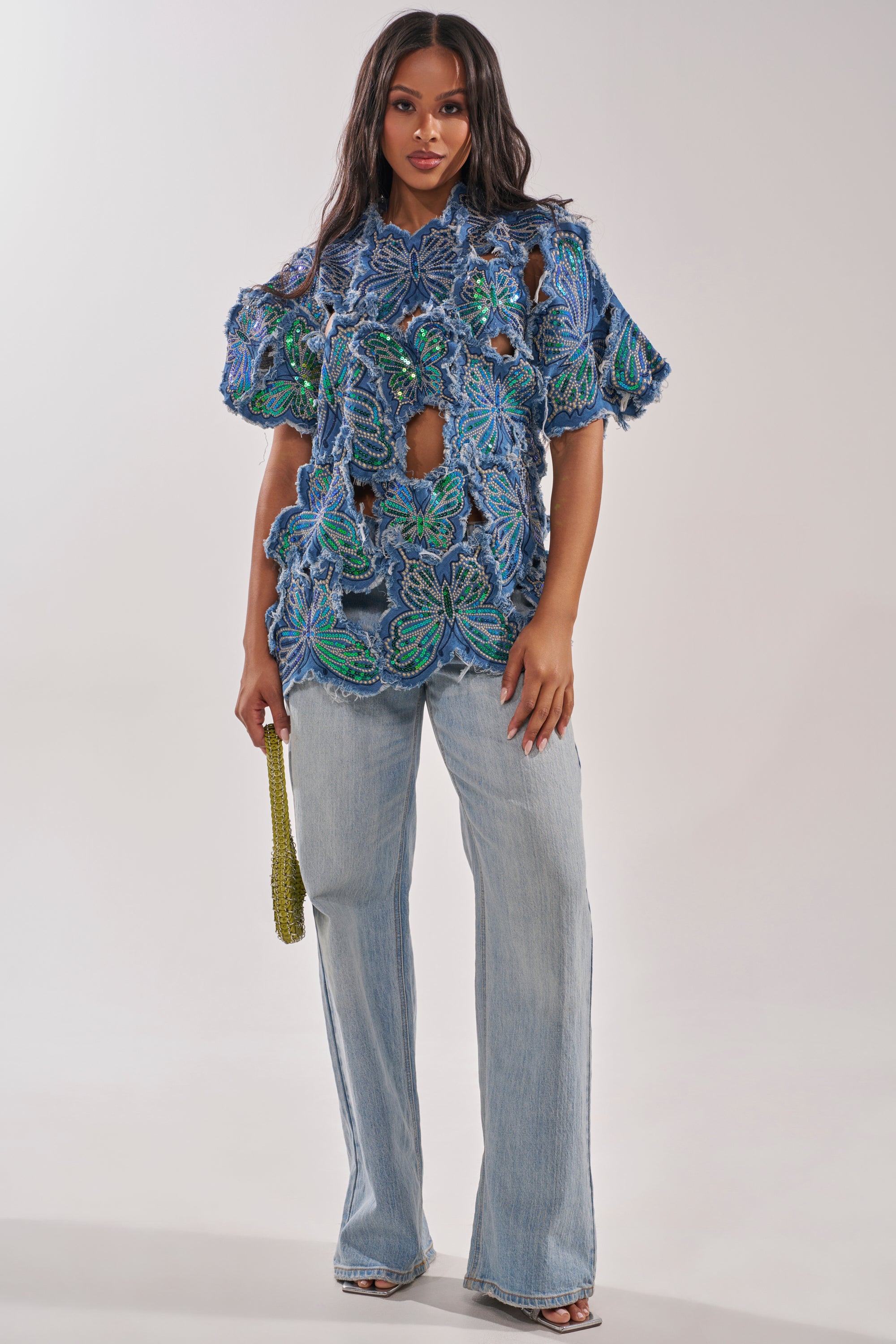 A woman faces forward in light blue wide-leg jeans and the SPREAD YOUR WINGS DENIM TOP, detailed with rhinestones, green leaf patterns, and cutouts. She carries a beaded green handbag and wears heeled sandals for a modern festival look. 5