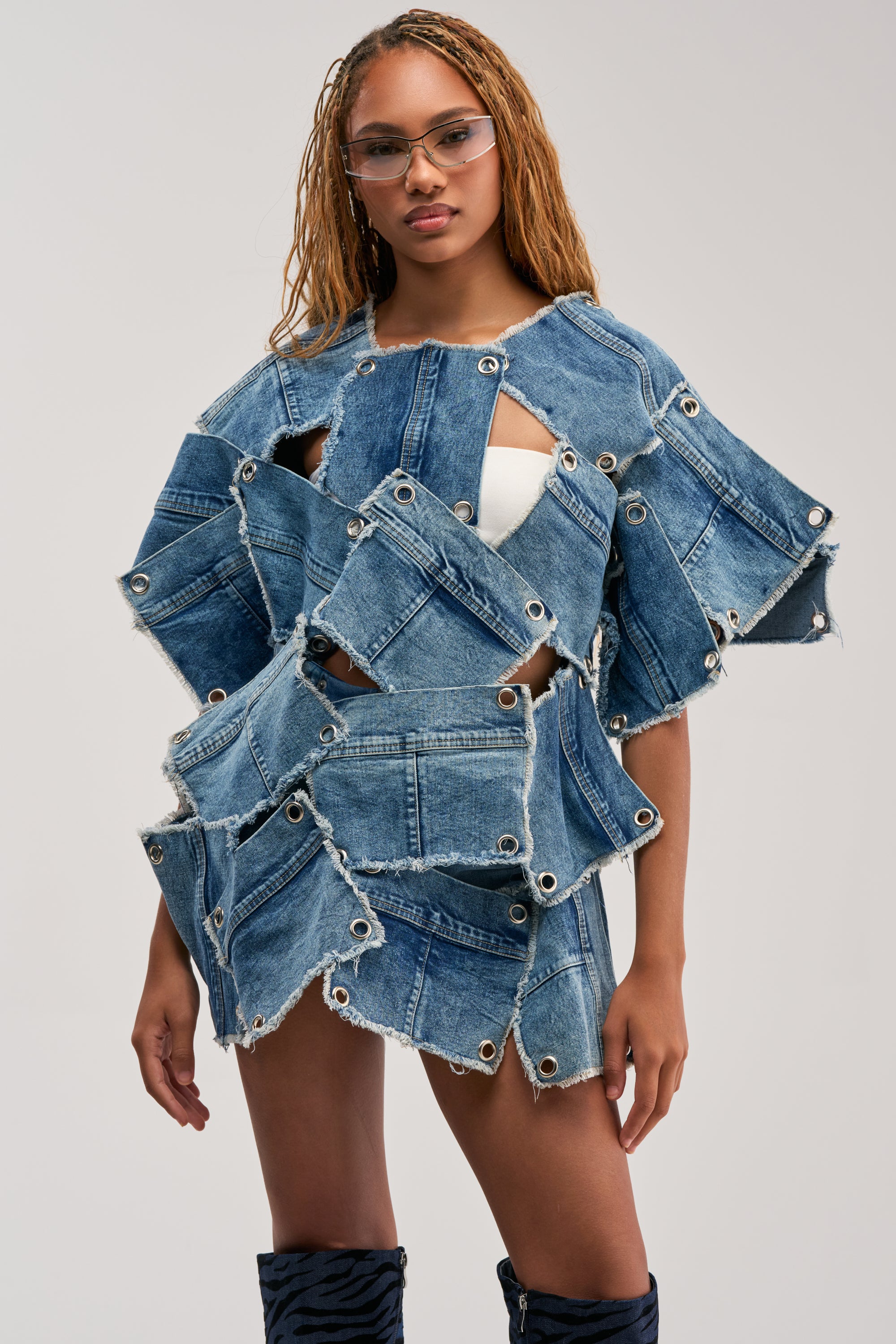 Wearing the ALL PATCHED UP DENIM TOP, which features overlapping distressed denim patches with frayed edges and metal grommets, a woman pairs it with a white skirt and clear glasses as she stands confidently against a neutral background.