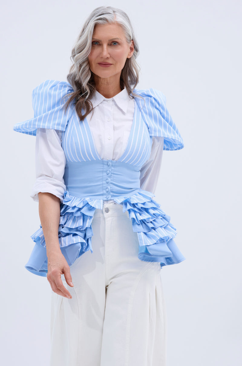 A woman with long, wavy gray hair wears the TIME OUT BLOUSE with puff sleeves and peplum design, styled with a matching corset-style vest and wide-legged white pants, standing against a plain light background.