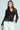 A woman with long brown hair is wearing a sheer black striped jacket over the SEDUCTION & BEAUTY STRUCTURED BLOUSE with white pants, and has a neutral expression with one hand in her pocket against a soft green backdrop, enhancing her Y2K-inspired look.