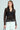 Someone with long hair is wearing a chic SEDUCTION & BEAUTY STRUCTURED BLOUSE, featuring sheer long sleeves over a black top. Paired with high-waisted white pants with buttons, they complete the Y2K-inspired look by holding a small black accessory.