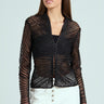 Someone with long hair is wearing a chic SEDUCTION & BEAUTY STRUCTURED BLOUSE, featuring sheer long sleeves over a black top. Paired with high-waisted white pants with buttons, they complete the Y2K-inspired look by holding a small black accessory.