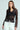 A person poses confidently in a SEDUCTION & BEAUTY STRUCTURED BLOUSE, featuring a sheer top over a black Y2K-inspired design and high-waisted white pants with buttons. They have long, straight hair and maintain a neutral expression against a light background.