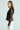 A person with long brown hair is wearing the SEDUCTION & BEAUTY STRUCTURED BLOUSE in black, paired with light-colored pants. They stand sideways, gaze at the camera against a plain background, radiating a Y2K-inspired look with diagonal stripes on the blouse.