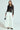 Dressed confidently in a Y2K-inspired look, a person flaunts a sheer SEDUCTION & BEAUTY STRUCTURED BLOUSE over a black bralette. They pair it with high-waisted white wide-leg trousers, black heels, and carry a small black handbag against the plain white background.