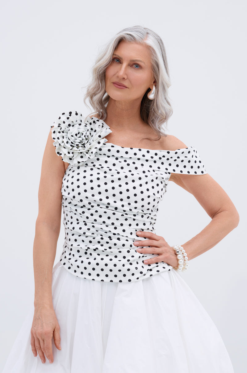 A woman with long gray hair wears the RUCHED POLKA DOT OFF THE SHOULDER TOP, which has black polka dots and a large fabric flower, paired with a white skirt. She stands confidently against a plain light background.