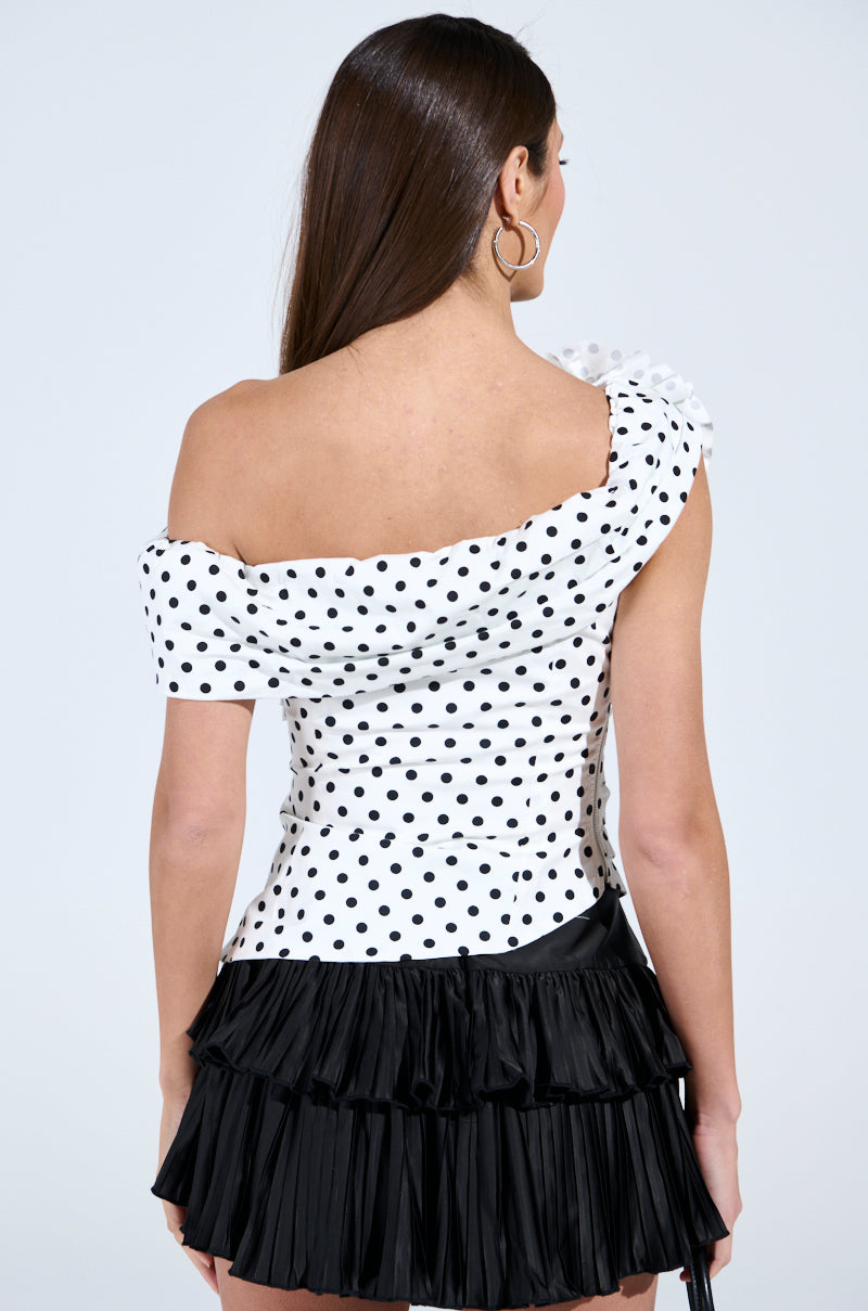 A woman with long brown hair poses against a light gray background, wearing the RUCHED POLKA DOT OFF THE SHOULDER TOP complemented by a short black ruffled skirt, her back facing the camera.