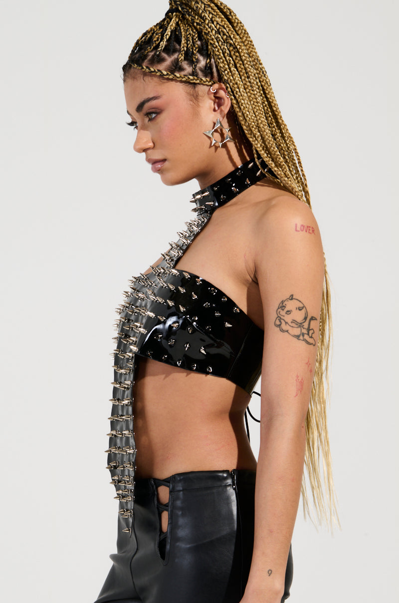 A person with long braided hair is wearing the GODS FAVORITE TOP, a black studded leather piece adorned with silver spike embellishments, and they are paired with matching patent faux leather pants. They accessorize with star earrings, and their tattoos are prominently visible, featuring a heart inscribed "LOVE" and a cartoon cat, all set against a plain white background. 4