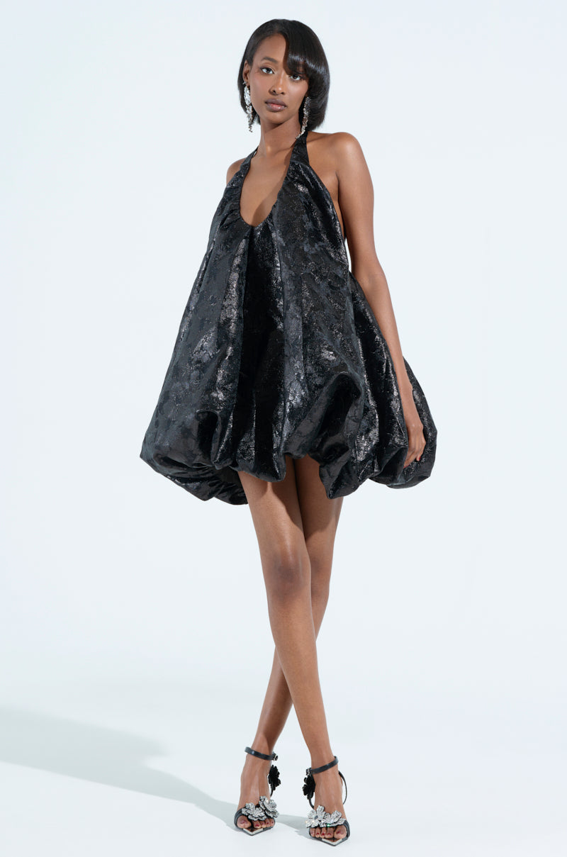 A woman poses confidently against a light background in an ON TREND BUBBLE BROCADE MINI DRESS, accentuated by black high-heeled sandals with decorative elements and large earrings. Her sleek hairstyle highlights the dress's elegant metallic fabrication.