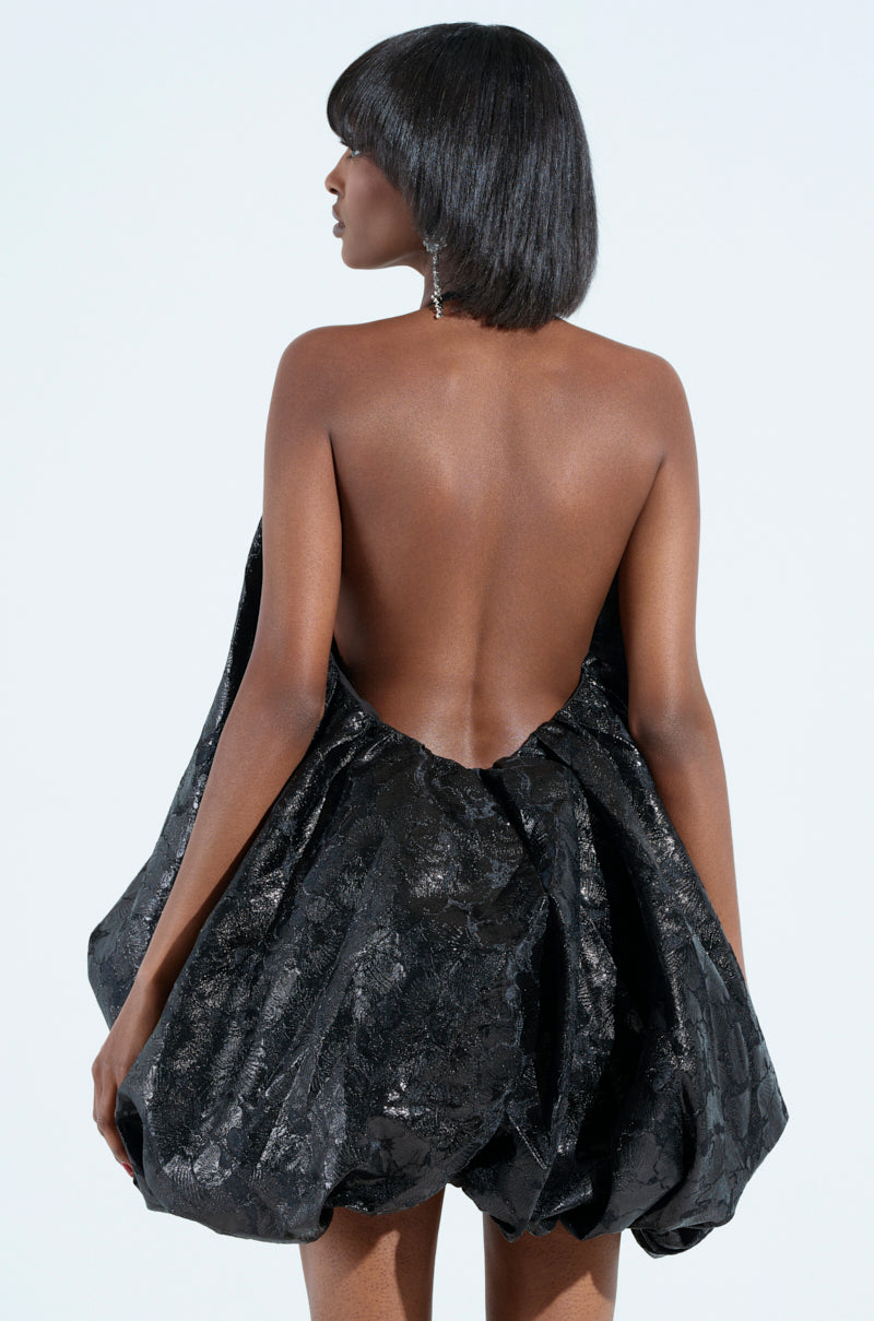 A woman with short dark hair stands against a plain background, facing away. She wears an ON TREND BUBBLE BROCADE MINI DRESS featuring a plunging V-neckline, open back, voluminous skirt, and textured pattern.