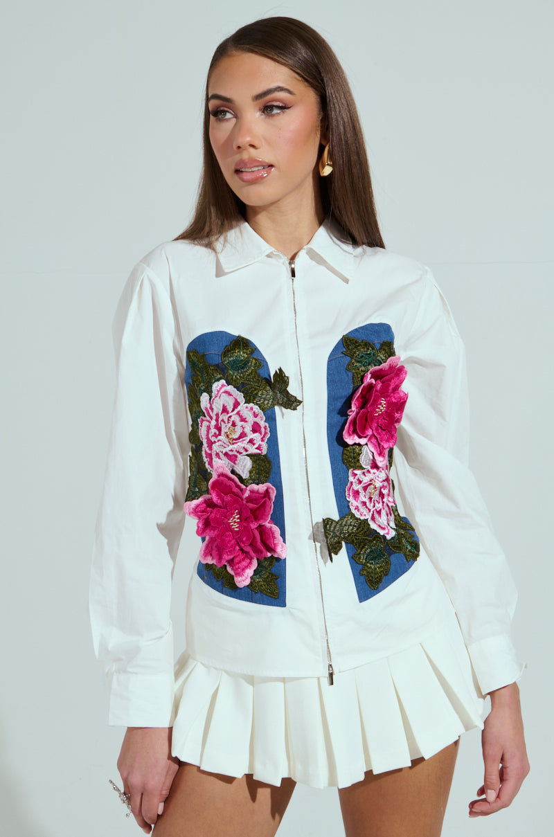 A woman with long brown hair wears a DAISY ZIPPER TOP featuring embroidered floral designs and blue panels, paired with a white pleated skirt, while standing against a plain background and looking to her left.