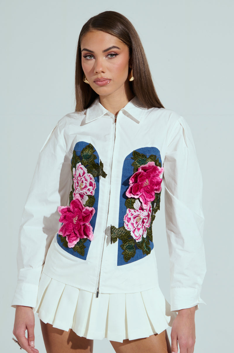 A woman in the DAISY ZIPPER TOP, a white long-sleeve button-up with pink floral embroidery, stands against a plain background. She pairs it with a white pleated skirt and gold circular earrings, her long brown hair cascading down.