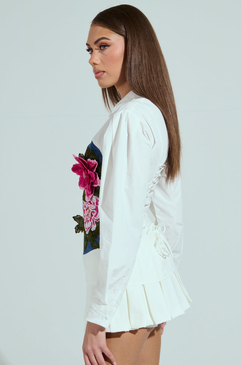 With long hair and side profile, a person wears the DAISY ZIPPER TOP showcasing exquisite pink and red embroidered floral graphics, paired elegantly with a short white pleated skirt against a plain background.
