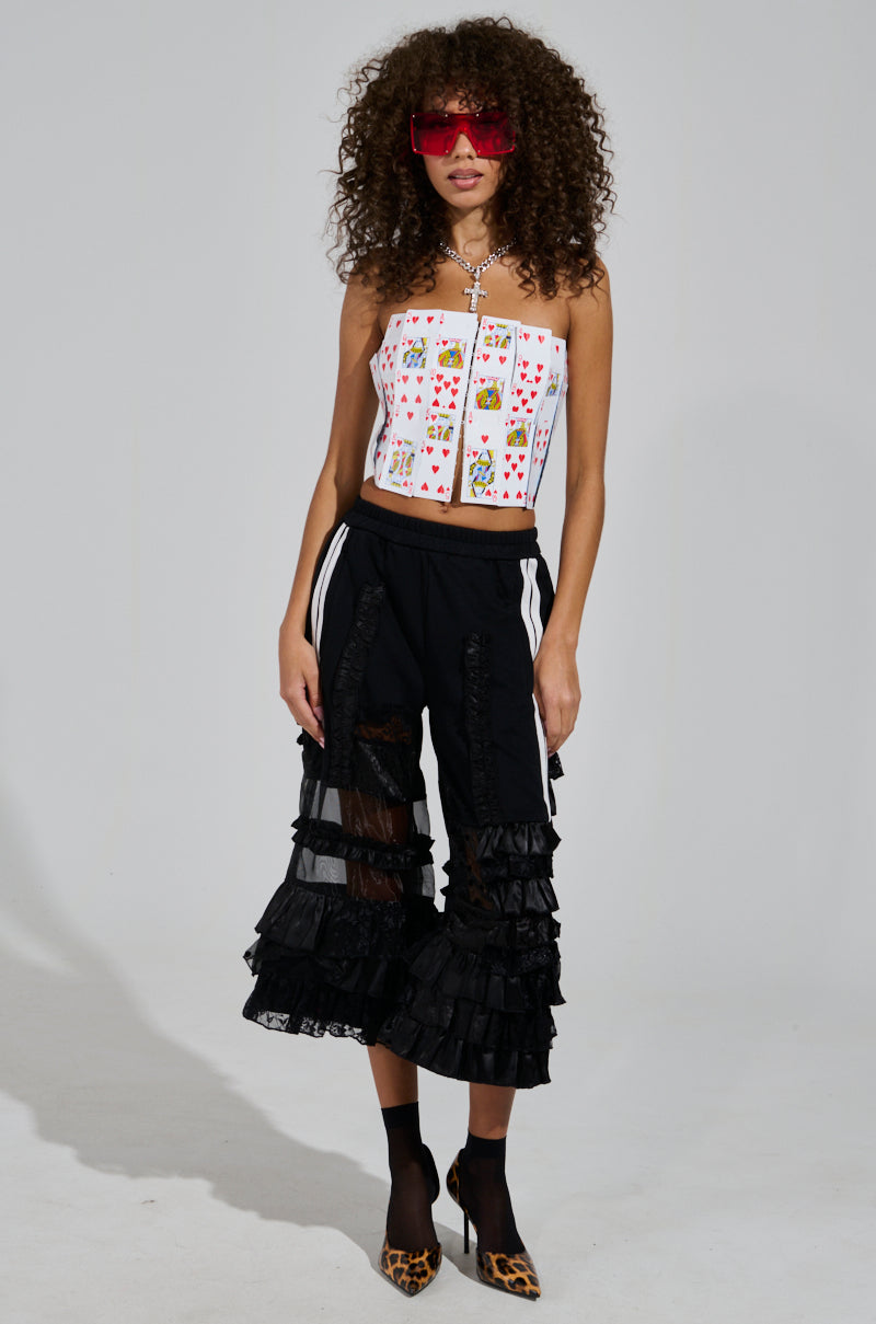 A person with curly hair wearing large red sunglasses, the LUCKY WINNER CORSET TOP featuring playing card appliques and a metallic back zipper, black ruffled pants, and leopard print shoes stands confidently against a plain background.