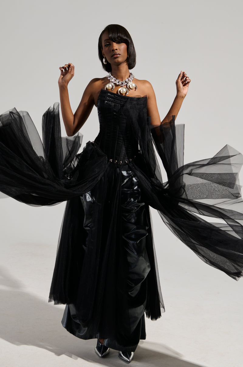 A person poses confidently in the MARRIED TO THE GAME Corset Top in black, featuring a flowing tulle skirt over a shiny satin base. A statement necklace adorns their neck, and their styled hair enhances the look. The ensemble is completed with a chic lace-up back, set against a plain background.