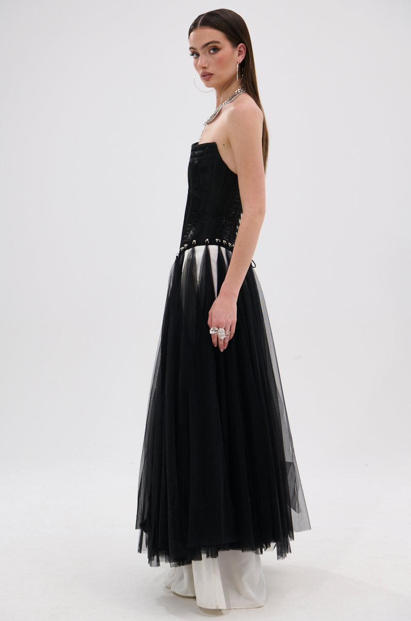 A woman in profile wears the MARRIED TO THE GAME CORSET TOP IN BLACK, a strapless piece with a lace-up back, styled with a long tulle skirt over white wide-leg pants. She has straight brown hair and layered jewelry, posing against a plain white background.