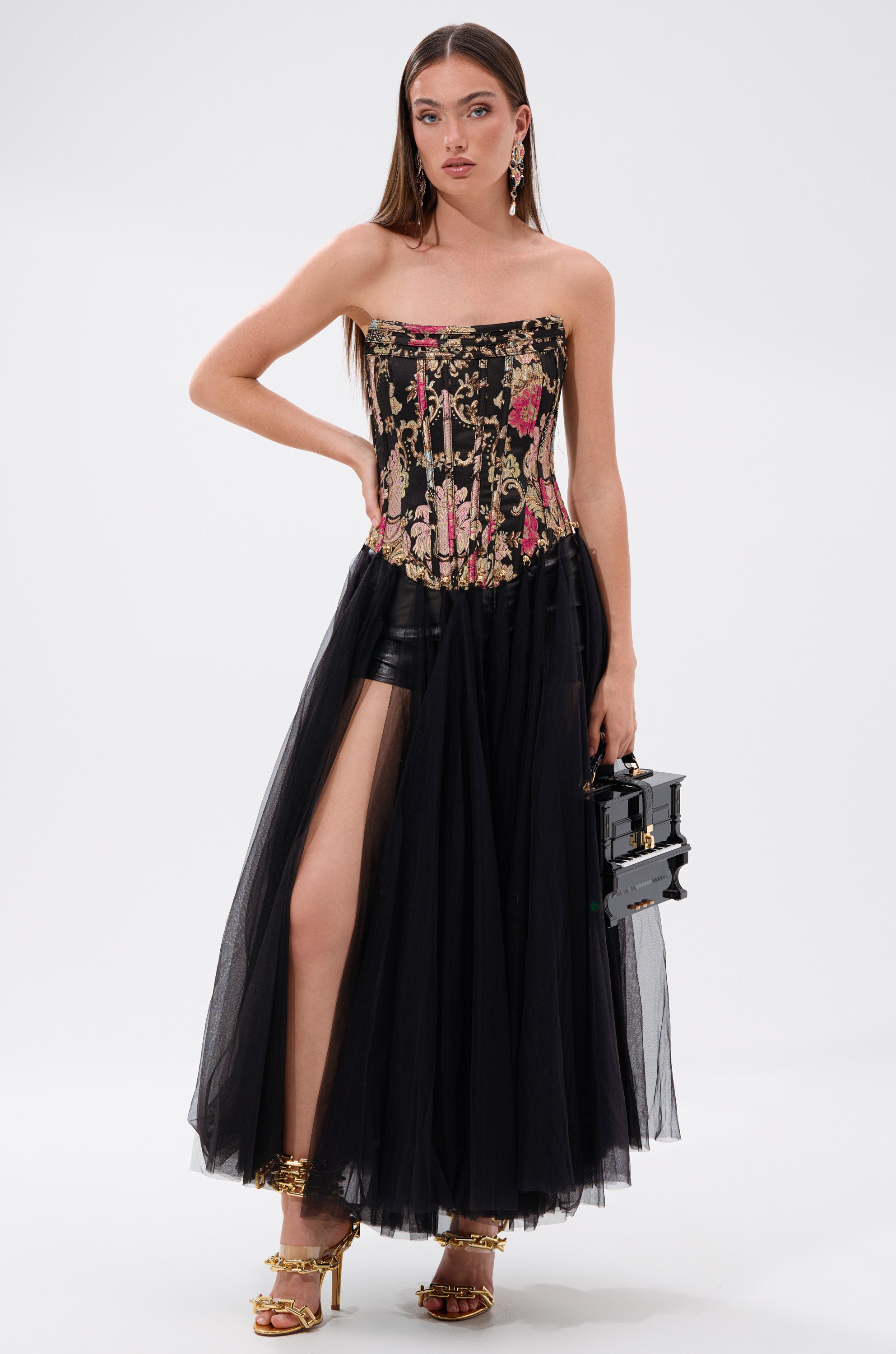 A woman models the ROYAL TULLE TRIM CORSET TOP with a long black tulle skirt featuring a high slit and black shorts. She accessorizes with a black handbag, gold high heels, and earrings against a plain white background. 0