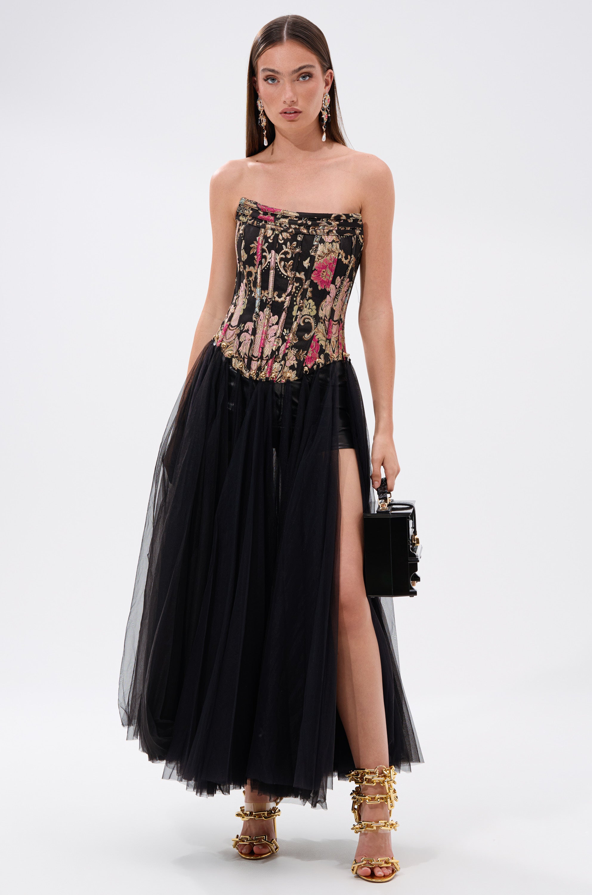 A woman wears the ROYAL TULLE TRIM CORSET TOP with a tulle skirt and high slit, accessorized with a black box handbag, gold high-heeled sandals, and statement earrings. 1