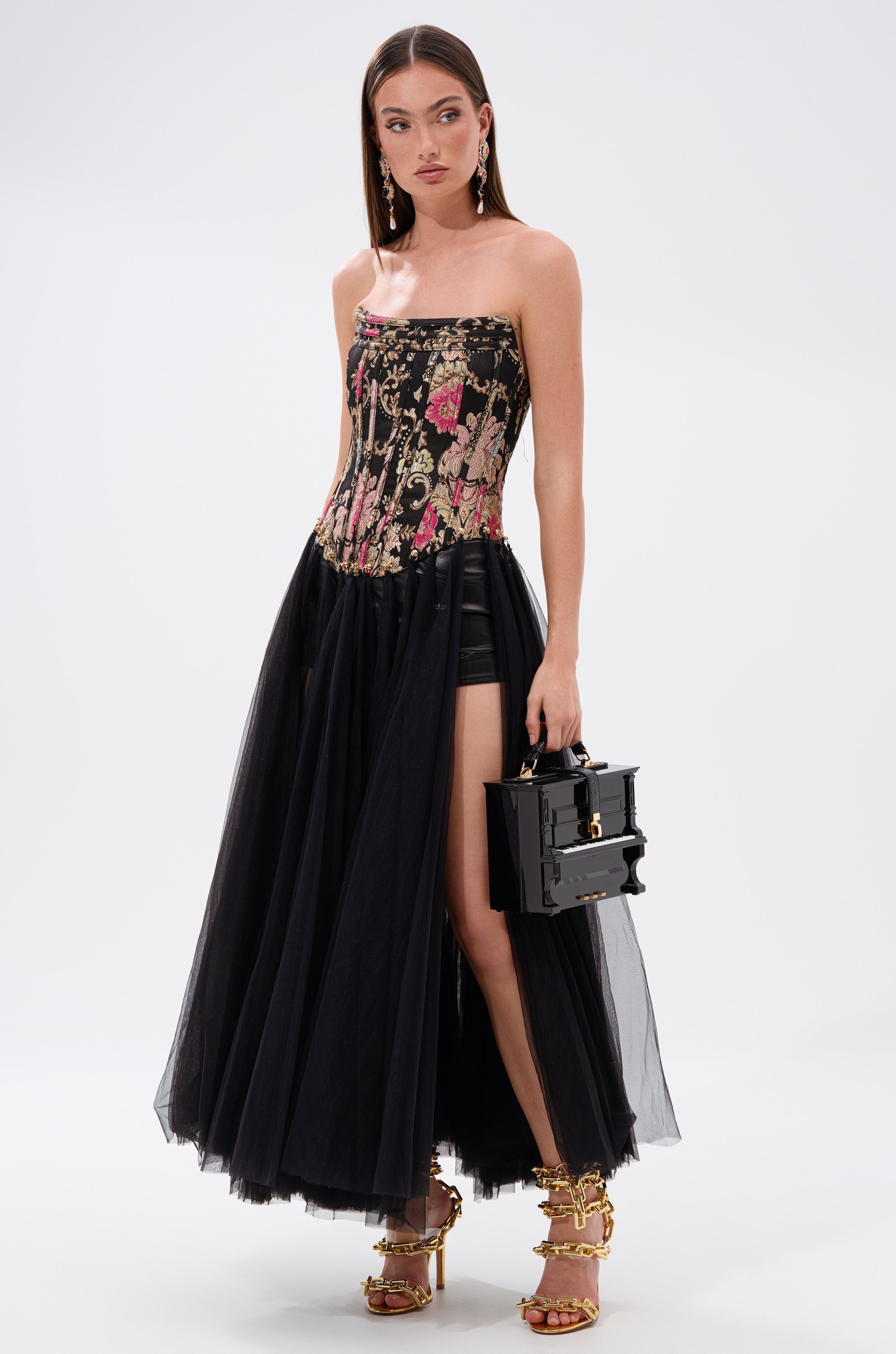 A woman wears the ROYAL TULLE TRIM CORSET TOP, a strapless black and gold floral design with tulle trim and sheer skirt featuring a thigh-high slit. She pairs it with gold heels, earrings, and a black handbag against a plain white background. 2