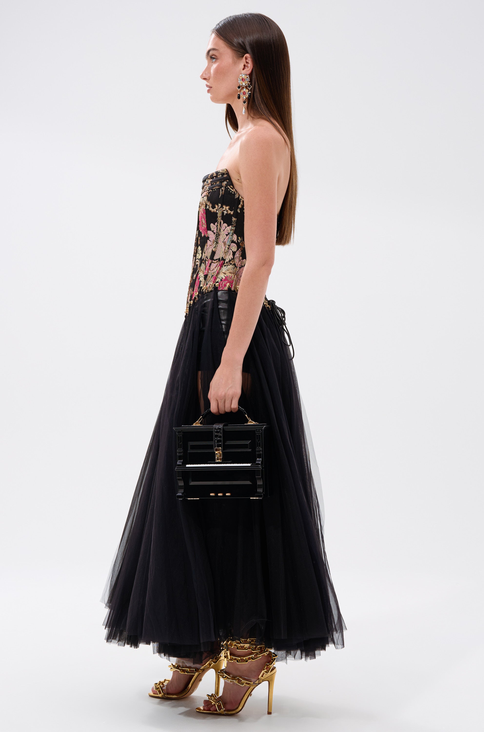 A woman in profile wears the ROYAL TULLE TRIM CORSET TOP with a strapless black tulle dress. She pairs it with a boxy handbag, gold high-heeled sandals, loose straight hair, and dangling earrings for a look of vintage elegance. 3