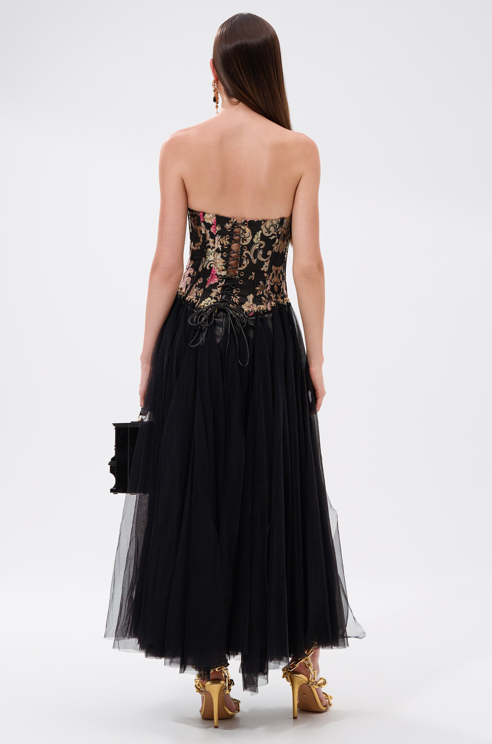A woman with straight brown hair wears the ROYAL TULLE TRIM CORSET TOP, a strapless floral corset, paired with a black tulle skirt. She carries a small black handbag and gold heels for a look of vintage elegance. 4
