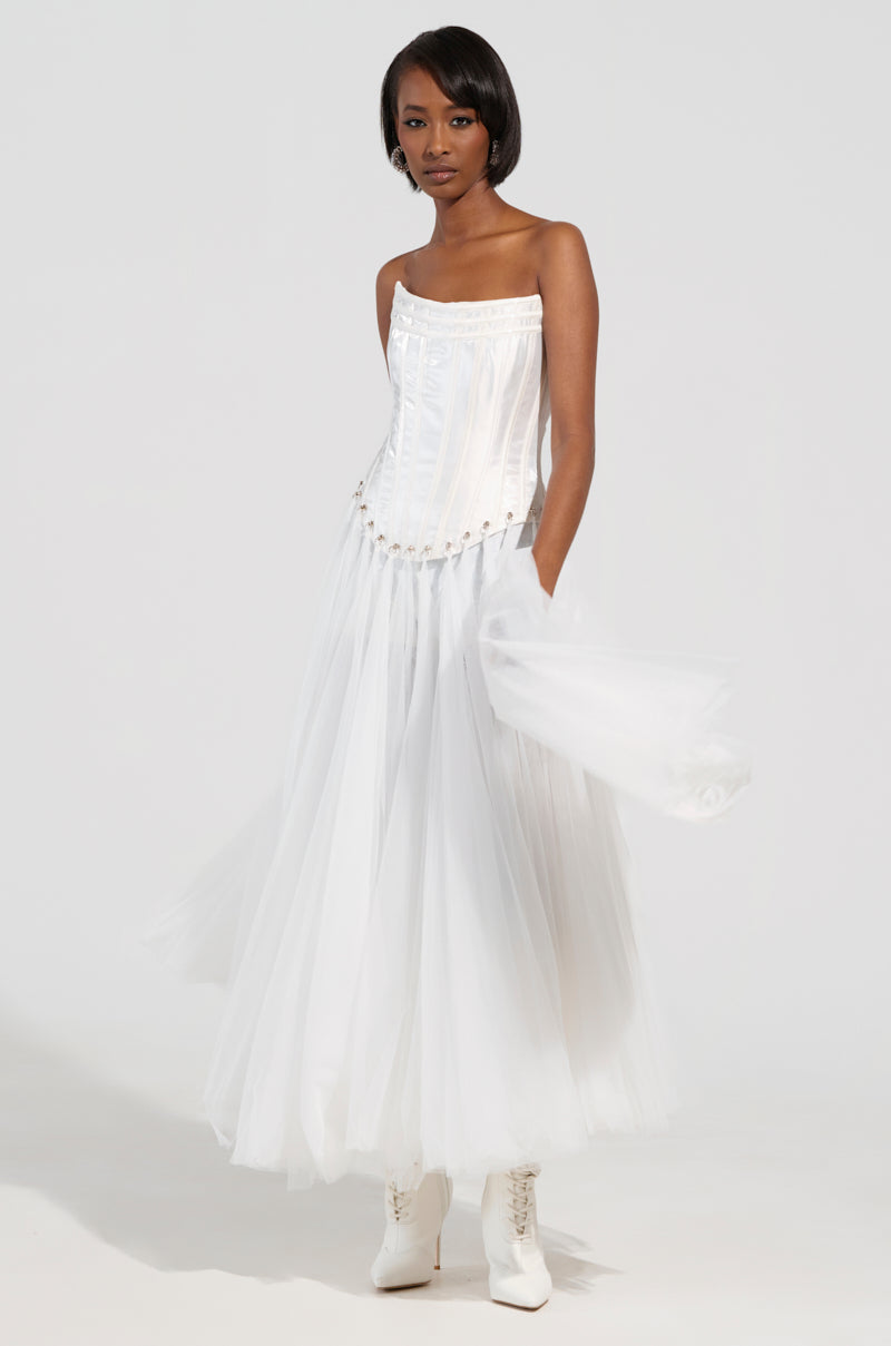 A person stands in the MARRIED TO THE GAME CORSET TOP IN WHITE, accompanied by a flowing tulle skirt. The outfit features a lace-up back panel, and they hold sheer white fabric in one hand while wearing white ankle boots. The backdrop is simple and white.