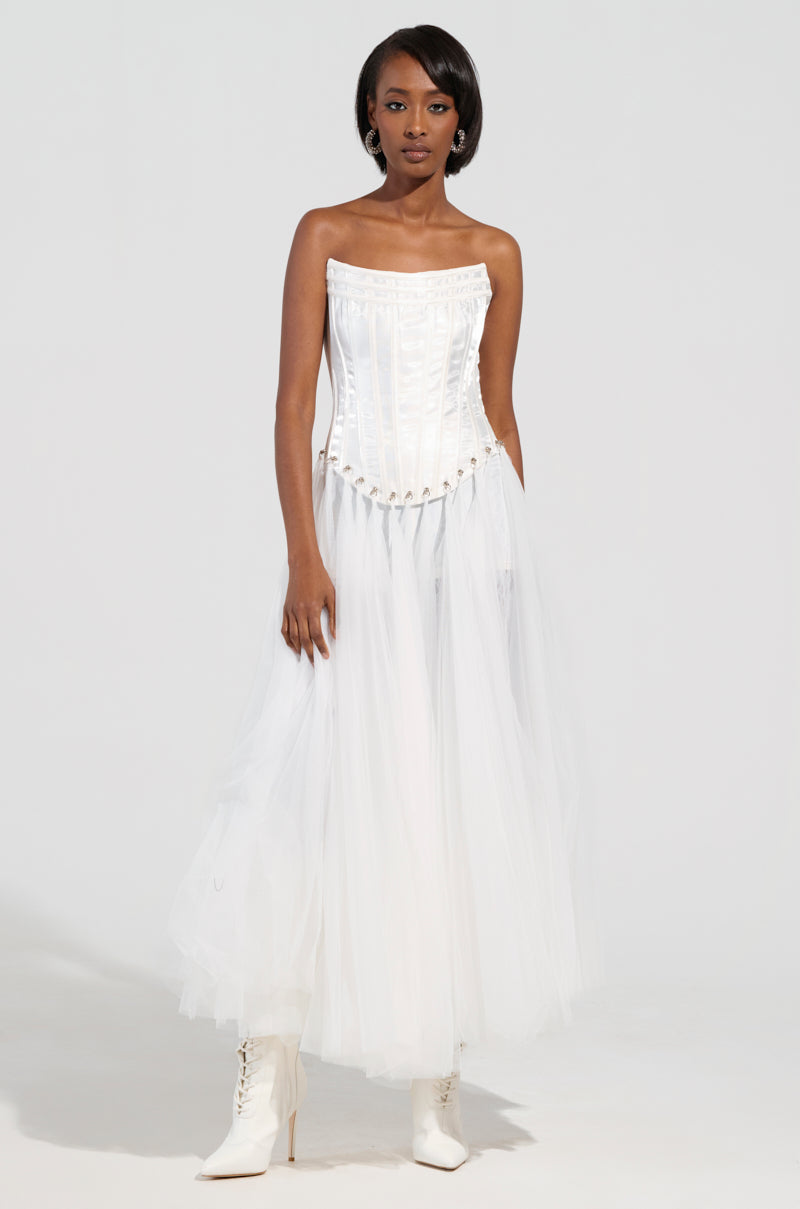 A woman poses gracefully against a plain light background, showcasing the "Married to the Game Corset Top in White," a strapless satin piece. She pairs it with a flowing tulle skirt and white ankle boots, highlighting its lace-up back design.