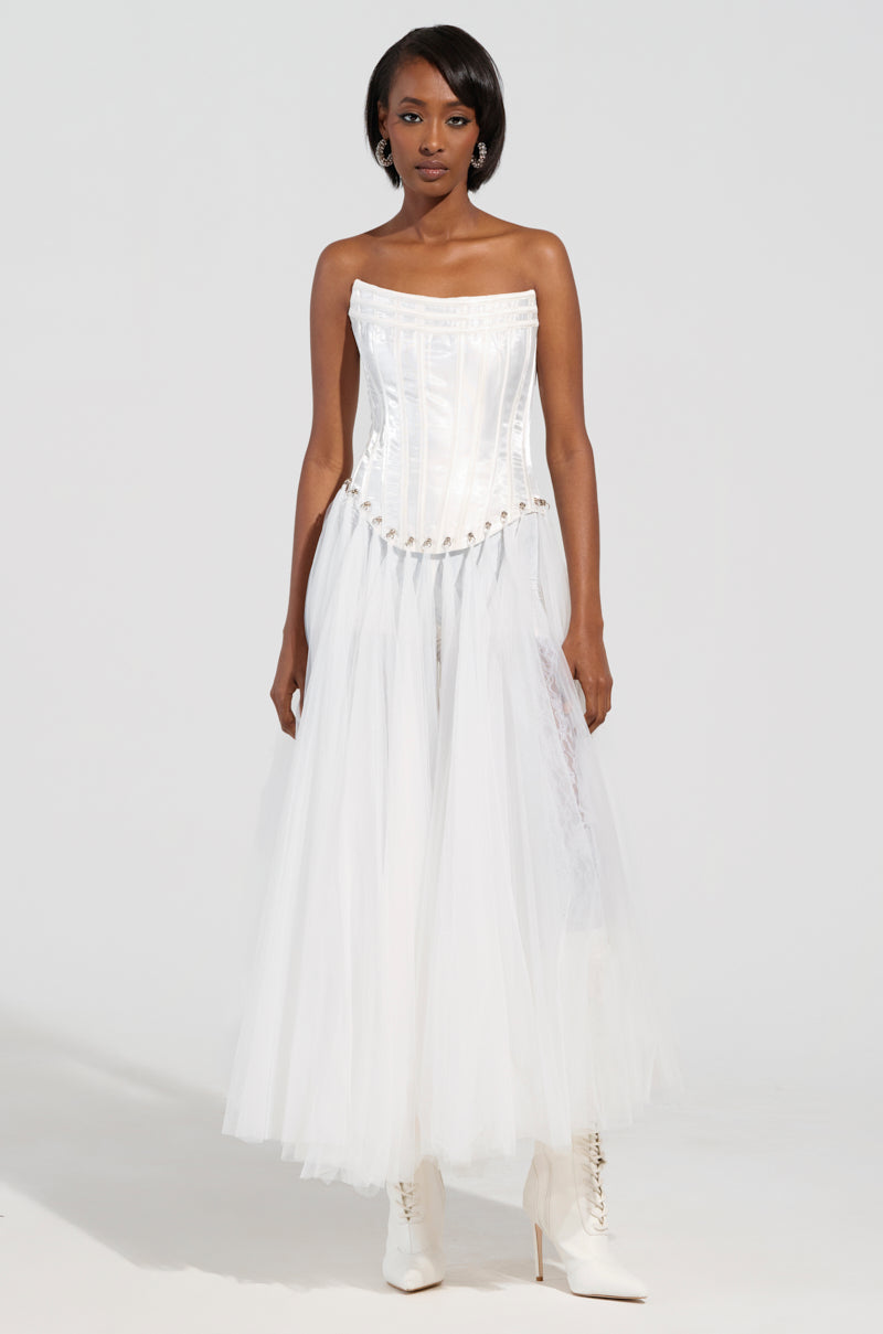 A person in a white "Married to the Game" corset top, featuring a lace-up back for added elegance, stands against a plain background. They pair it with a tulle skirt and white ankle boots.
