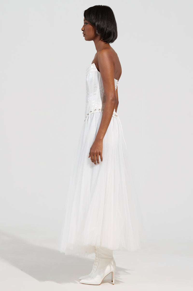 A woman stands in profile wearing the "Married to the Game Corset Top in White," featuring a strapless satin design with a tulle skirt and a waist-cinching bodice, paired with white high-heeled boots. The background is plain and neutral.