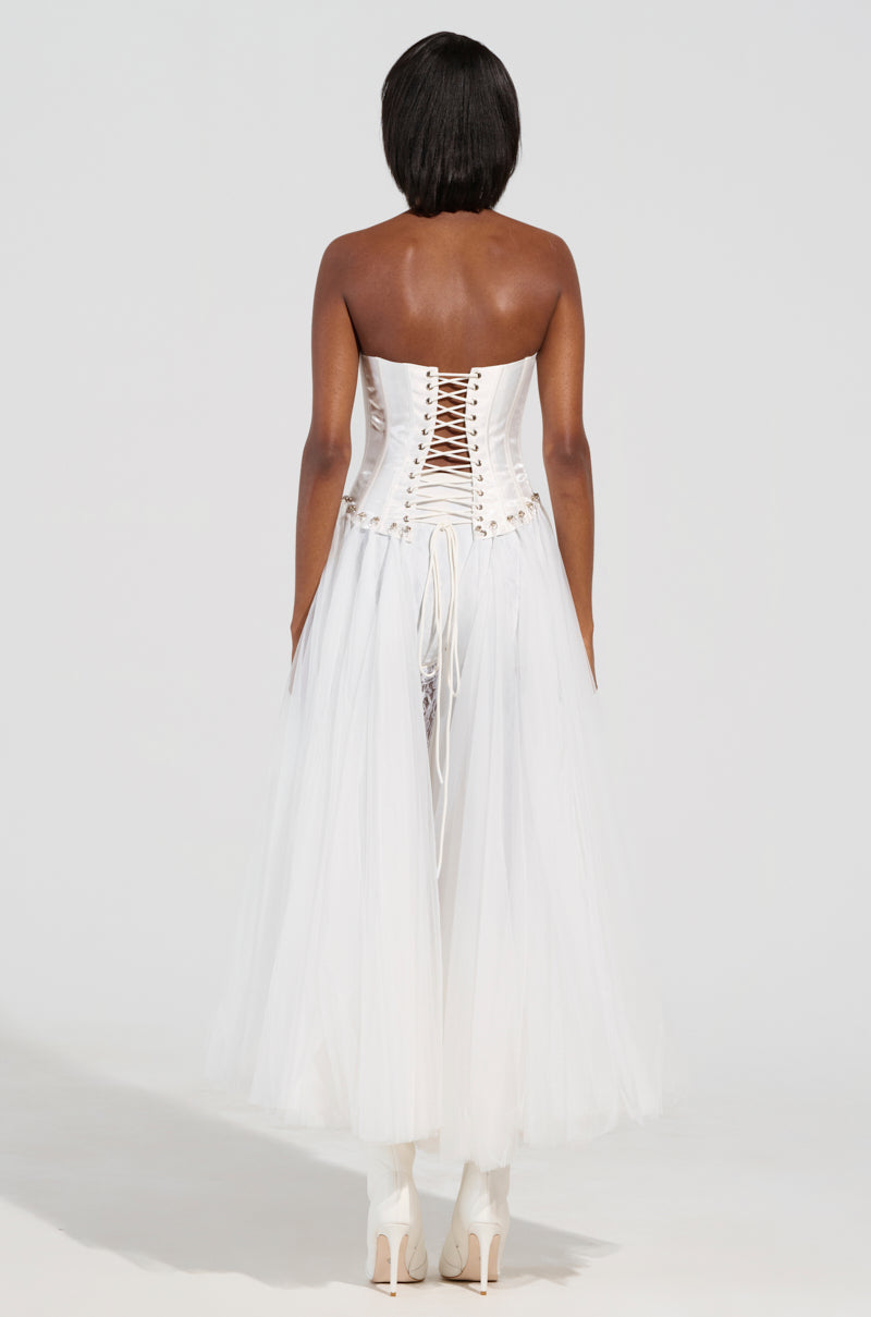 A person wearing the "Married to the Game" corset top in white, featuring intricate lacing and paired with a flowing tulle skirt, stands with their back facing the camera. They complete their outfit with high-heeled shoes.
