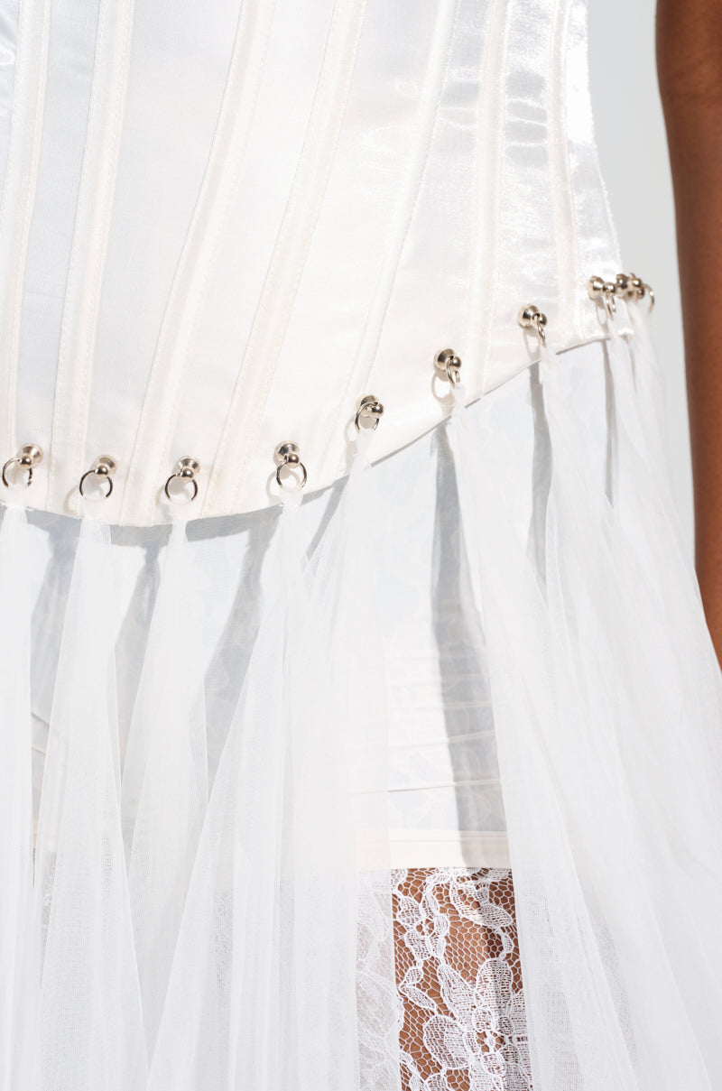 A close-up of the "Married to the Game Corset Top in White" showcases its strapless satin design, highlighted by metal ring accents and sheer white lace fabric that gracefully hangs below. The corset's shimmering texture is complemented by a delicate lace-up back.