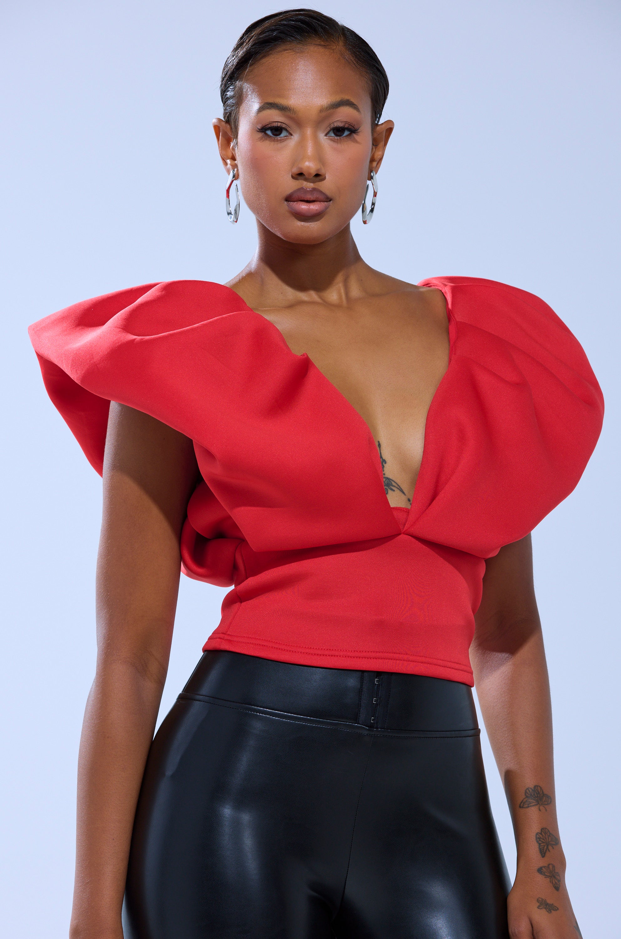A woman with short, slicked-back hair wears the FEMME ENERGY SCUBA BLOUSE IN RED with black shiny pants, hoop earrings, and visible tattoos as she poses against a light background.