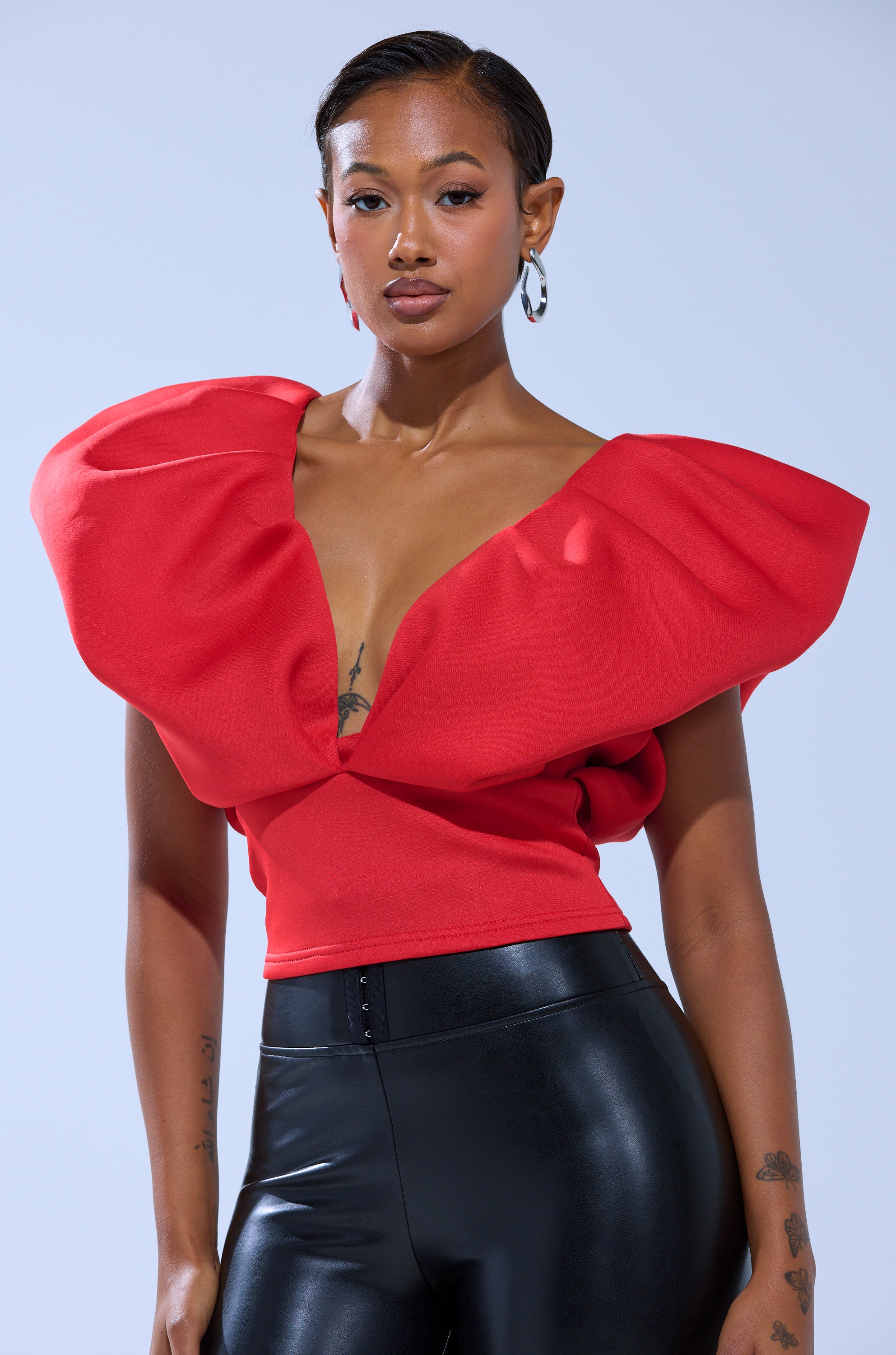 A woman models the FEMME ENERGY SCUBA BLOUSE IN RED, featuring pleated puff shoulders, paired with black shiny high-waisted pants. She poses against a light background, highlighting her short hair, hoop earrings, and arm tattoos.