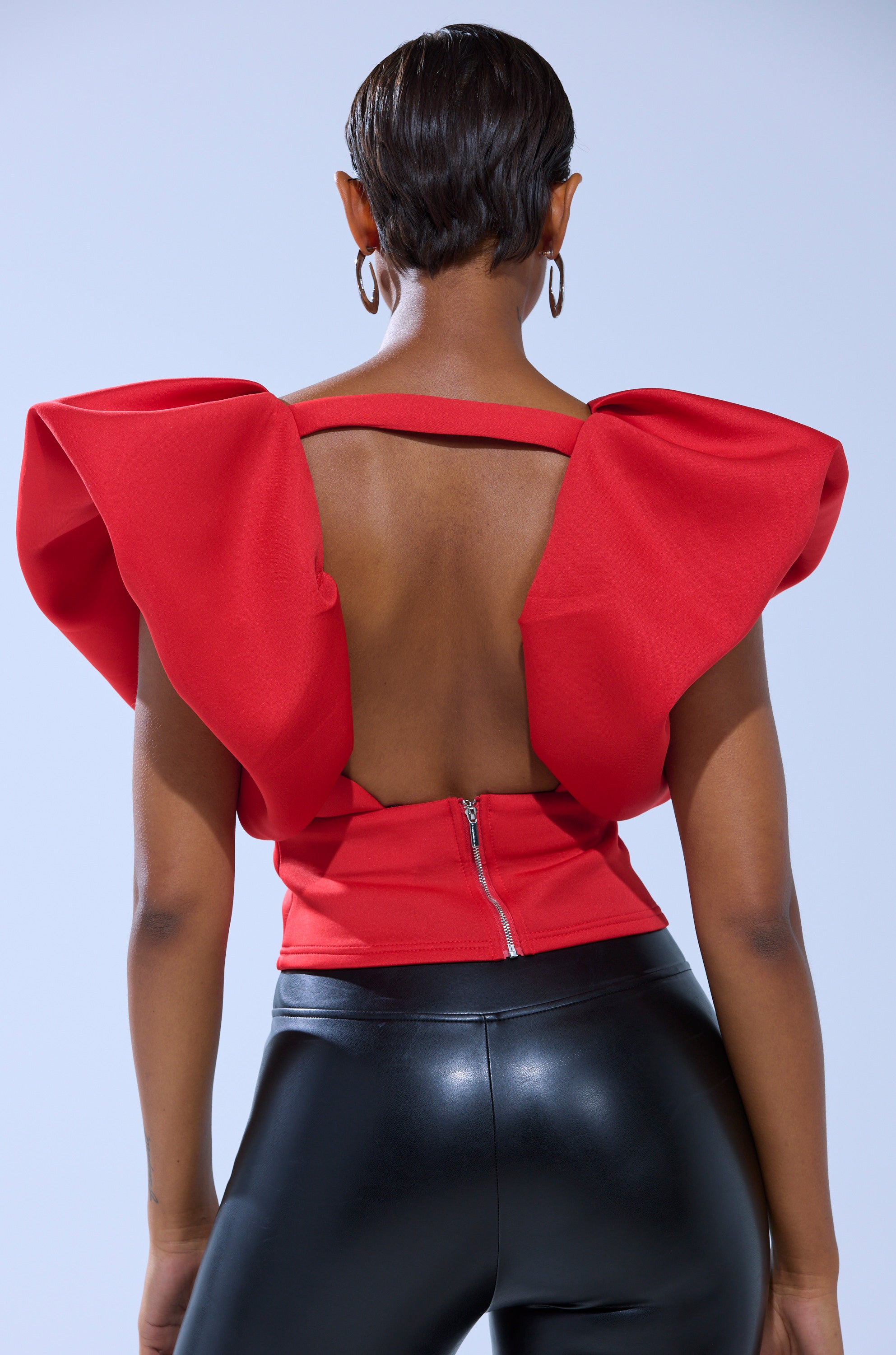 A person is shown from behind wearing the FEMME ENERGY SCUBA BLOUSE IN RED, featuring dramatic puff shoulder ruffled sleeves, paired with shiny black pants against a plain background.