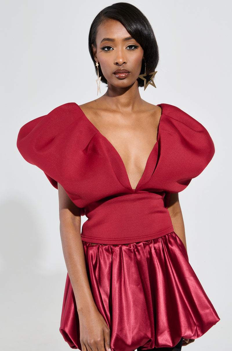 A woman in a bold burgundy dress, inspired by the Femme Energy Scuba Blouse, with dramatic puffed sleeves and a plunging V-neckline stands confidently. Her sleek, shoulder-length hair complements the star-shaped earrings she wears. The dress showcases a fitted bodice that flares out into a voluminous skirt.