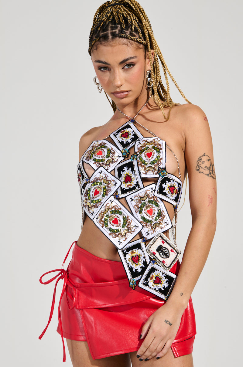 A person with braided hair strikes a pose in the QUEEN OF HEARTS TOP, an asymmetrical novelty piece crafted from playing cards, paired with a red leather skirt featuring ties. Hoop earrings dangle as their tattooed arm adds flair against the plain white background, completing this striking ensemble.