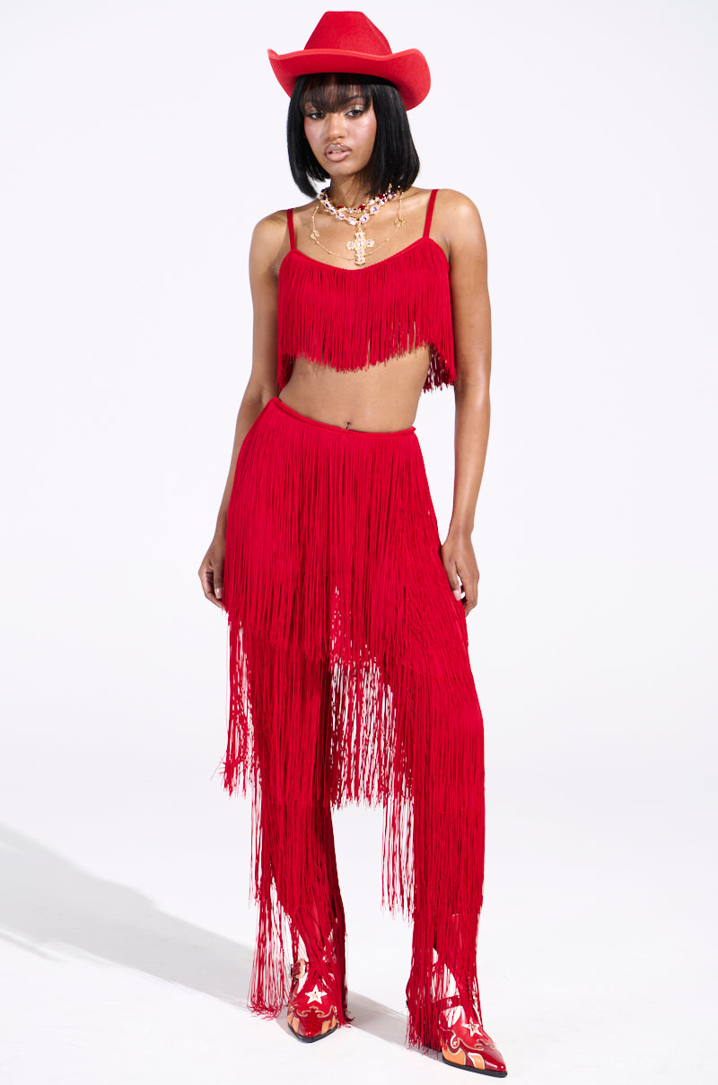 Wearing vibrant OH SO FRINGE PANTS IN RED with a coordinating fringed top, red cowboy hat, boots, and a gold necklace, this ensemble pops against the white background. The high-rise fit adds a stylish touch to the overall look.
