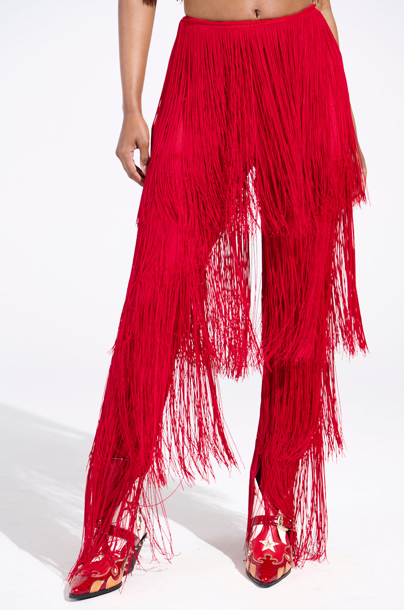 Dressed in OH SO FRINGE PANTS IN RED, a person stands against a white background, complementing the pants' vibrant long strands with matching lustrous red star-patterned shoes.