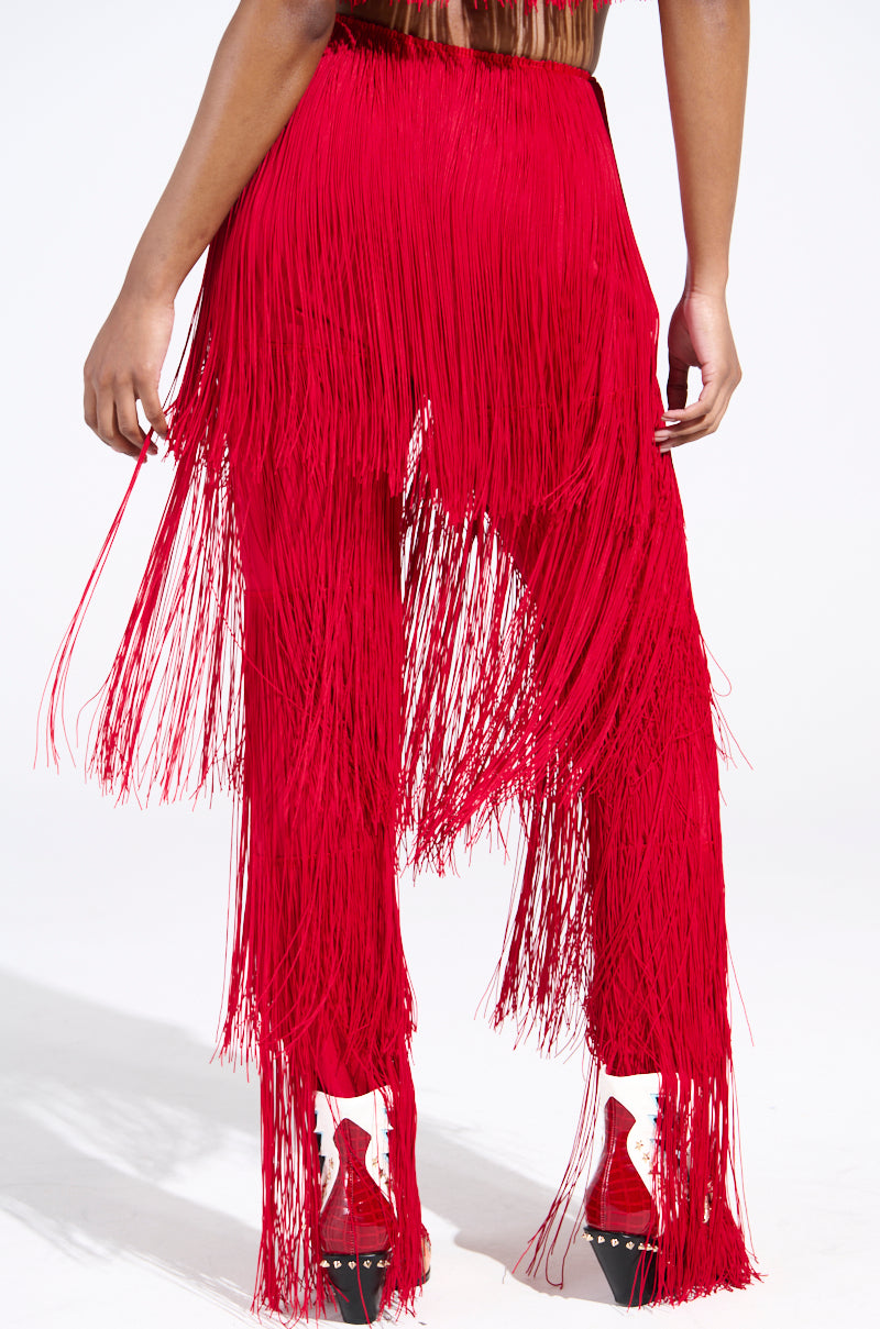 A person showcases the OH SO FRINGE PANTS IN RED, high-rise vibrant fringe pants paired with high heels. The dynamic, flowing fringes and plain background accentuate this bold fashion statement, crafted from stretchy fabric for ultimate comfort.