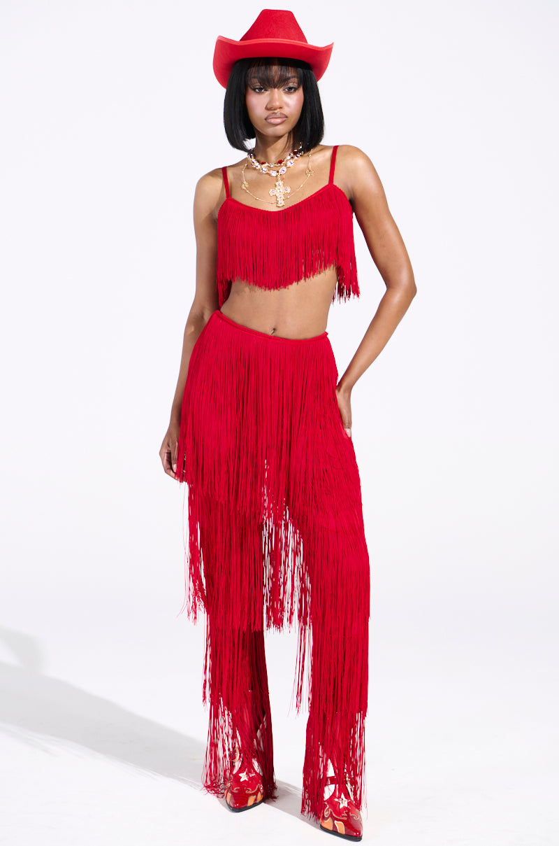 A person poses confidently against a plain white background, wearing OH SO FRINGE PANTS IN RED paired with a crop top and high-rise fit, accessorized with a red cowboy hat and necklace.