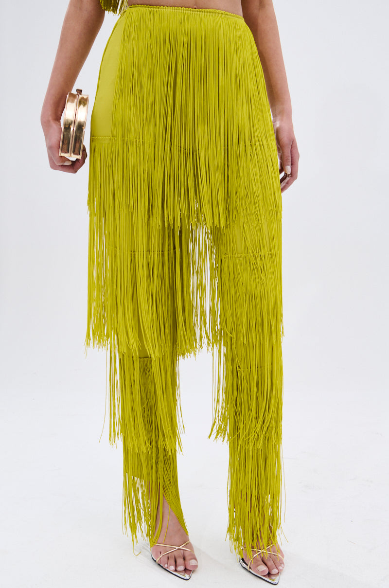 The lower half of a person wearing OH SO FRINGE PANTS IN GREEN, high-rise and stretchy with fringe detail, paired with open-toe high heel sandals and holding a small gold clutch. Matching top not visible.