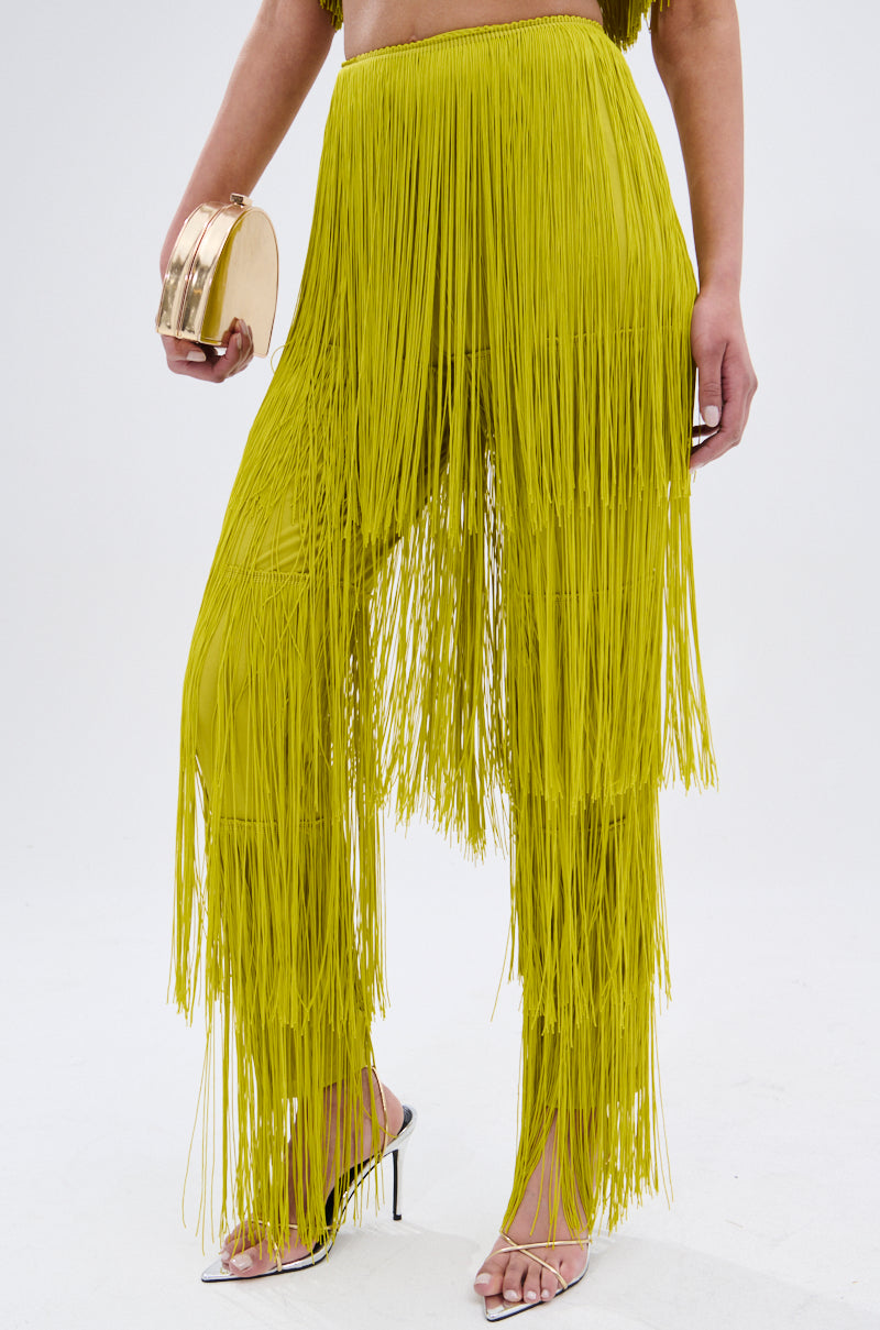 A person wearing OH SO FRINGE PANTS IN GREEN, featuring a high-rise fit and stretchy fabric, holds a round gold clutch and wears clear high-heeled sandals while standing against a plain light background.