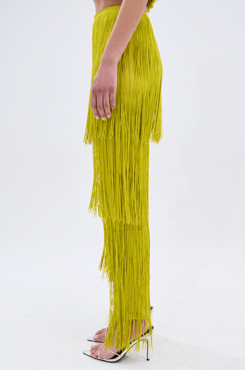 A person stands sideways in OH SO FRINGE PANTS IN GREEN, made from stretchy fabric, with clear high-heeled shoes. Only the lower half of their body is visible against a plain white background.